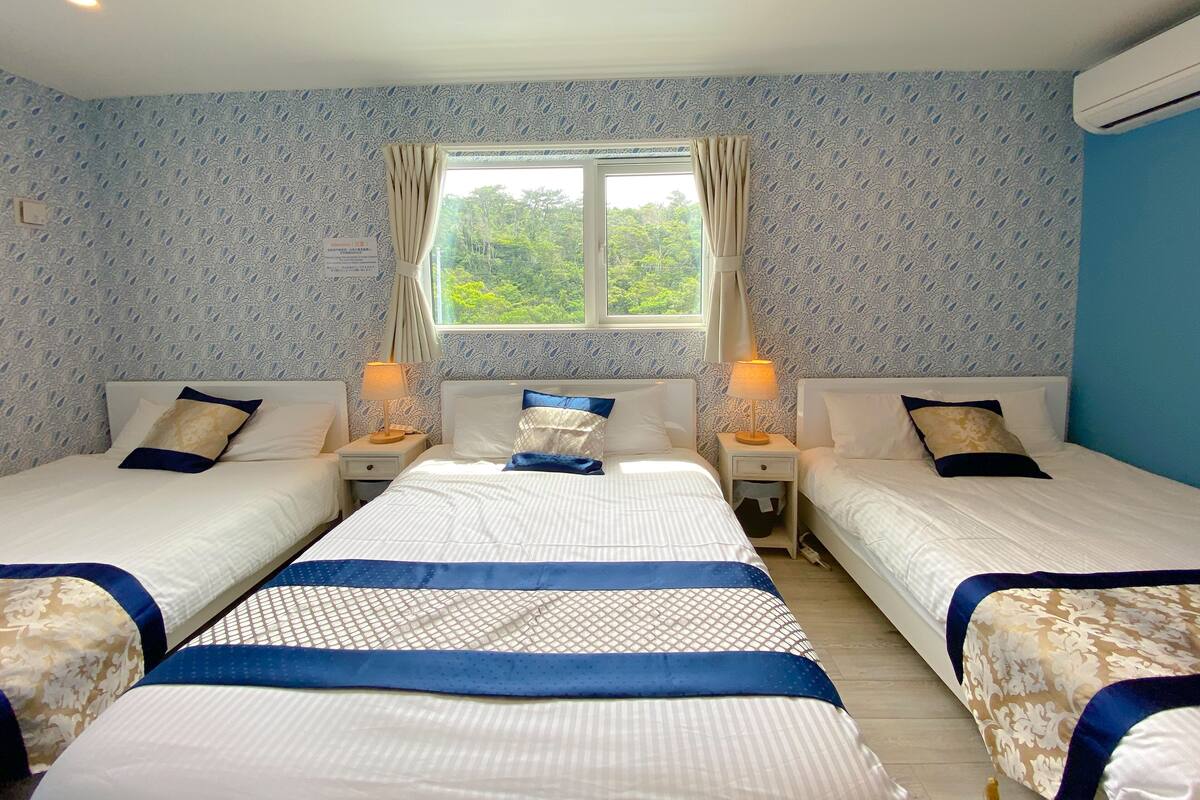 A hotel room with two beds, a window with curtains, patterned wallpaper, and a ceiling light.