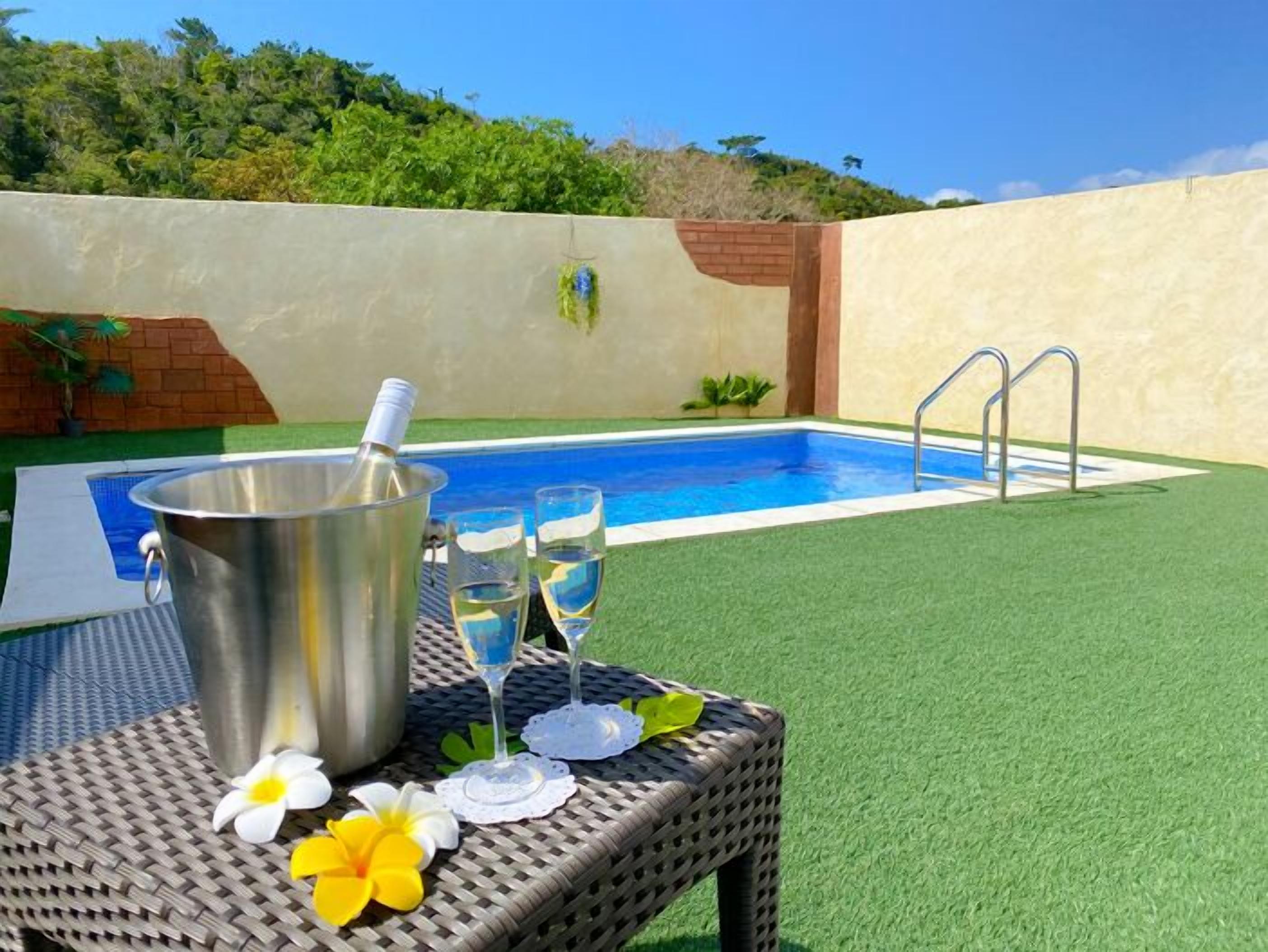 Une piscine avec un échelle en métal, un seau et deux verres d’eau sur une table en osier décorée de fleurs.