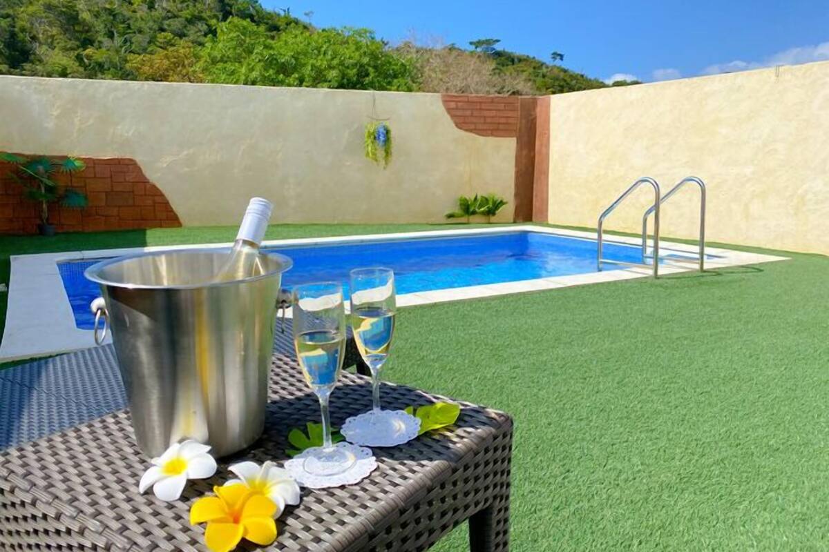 A swimming pool with a metal ladder, a bucket, and two glasses of water on a wicker table with flowers.