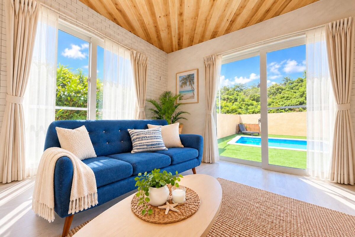 A modern living room with a blue sofa, a coffee table with a plant and a glass, and a view of a swimming pool through large windows.