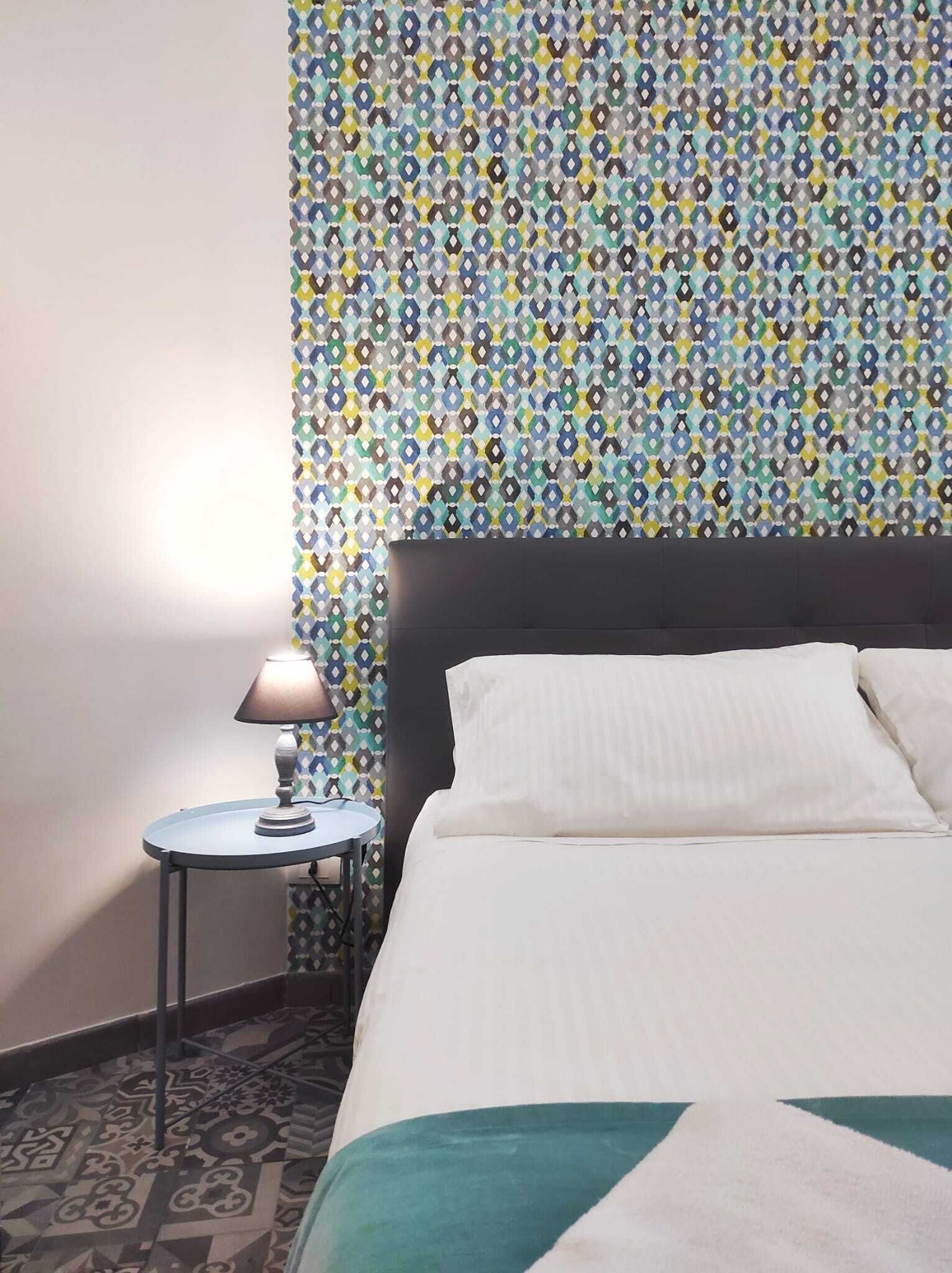 Una habitación de hotel con papel tapiz estampado, una mesita redonda con lámpara y una cama con ropa blanca.