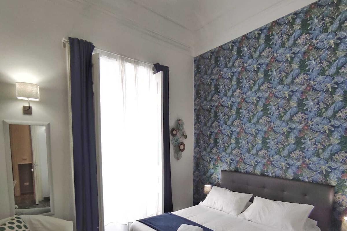 Une chambre d’hôtel avec un lit, une chaise, une table sur laquelle se trouve un livre et un mur décoré d’un motif.