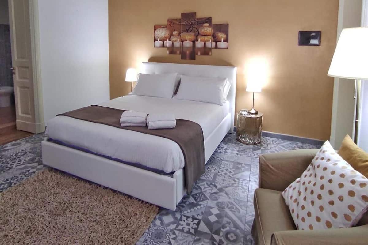 Une chambre d’hôtel avec un grand lit, une tête de lit ornée d’un panneau décoratif, une table de chevet avec une lampe, un tapis à motifs et un canapé agrémenté d’un coussin à pois.