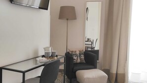 Habitación de hotel con televisor montado en la pared, escritorio con silla, lámpara, sofá con reposapiés y piso estampado.