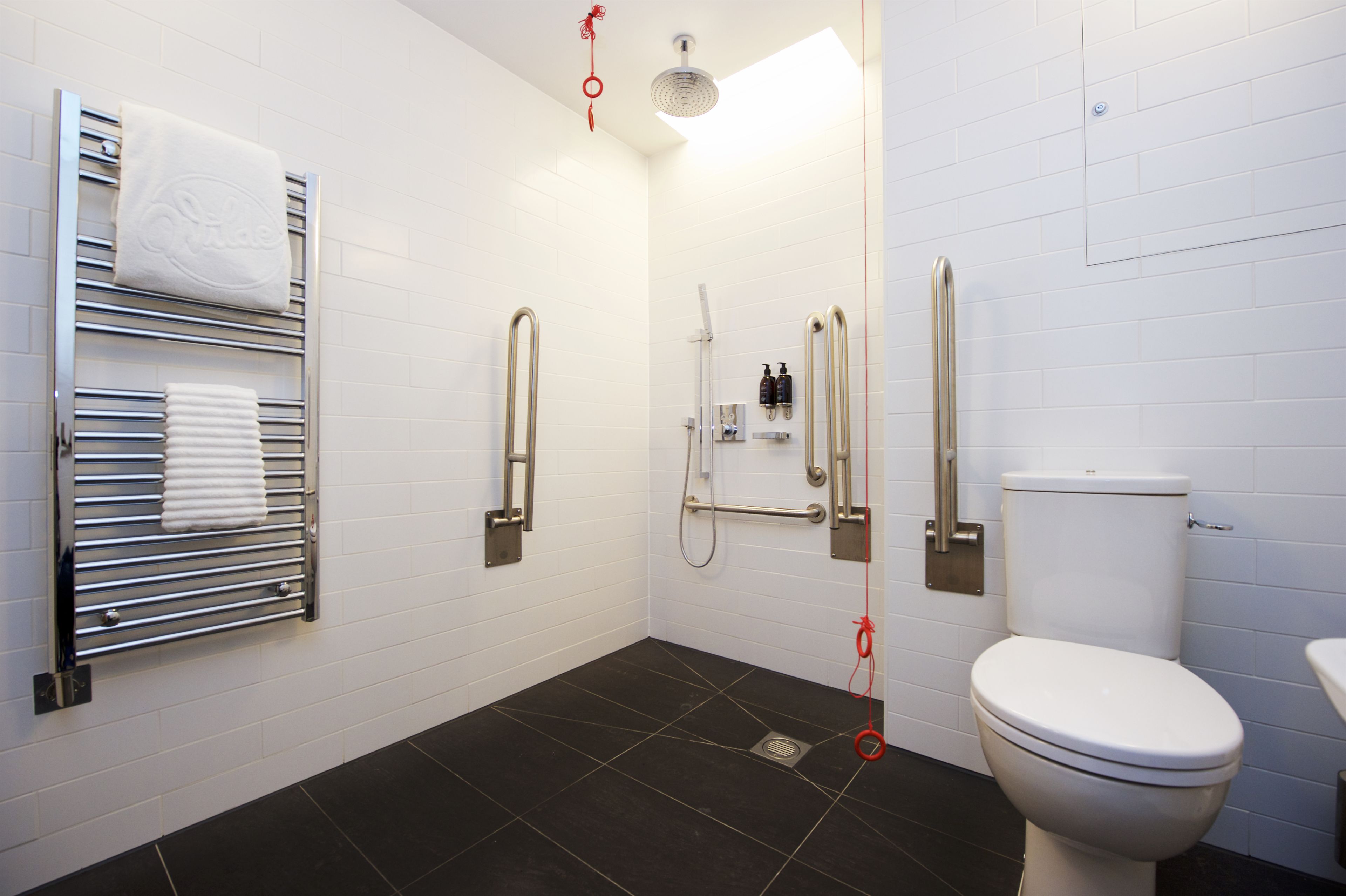 A modern bathroom with a white toilet, a shower with a handheld showerhead, a towel rack with folded towels, and a handrail.