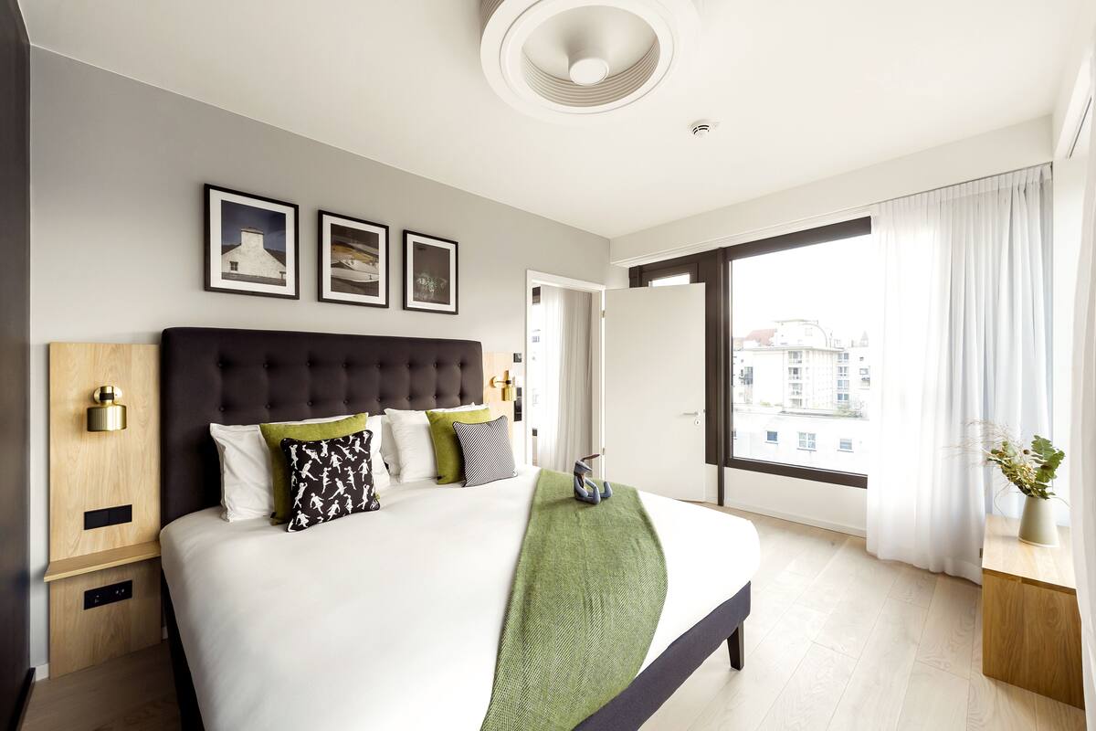 A modern hotel room with a large bed, a headboard, a bedside table, and a window with curtains.