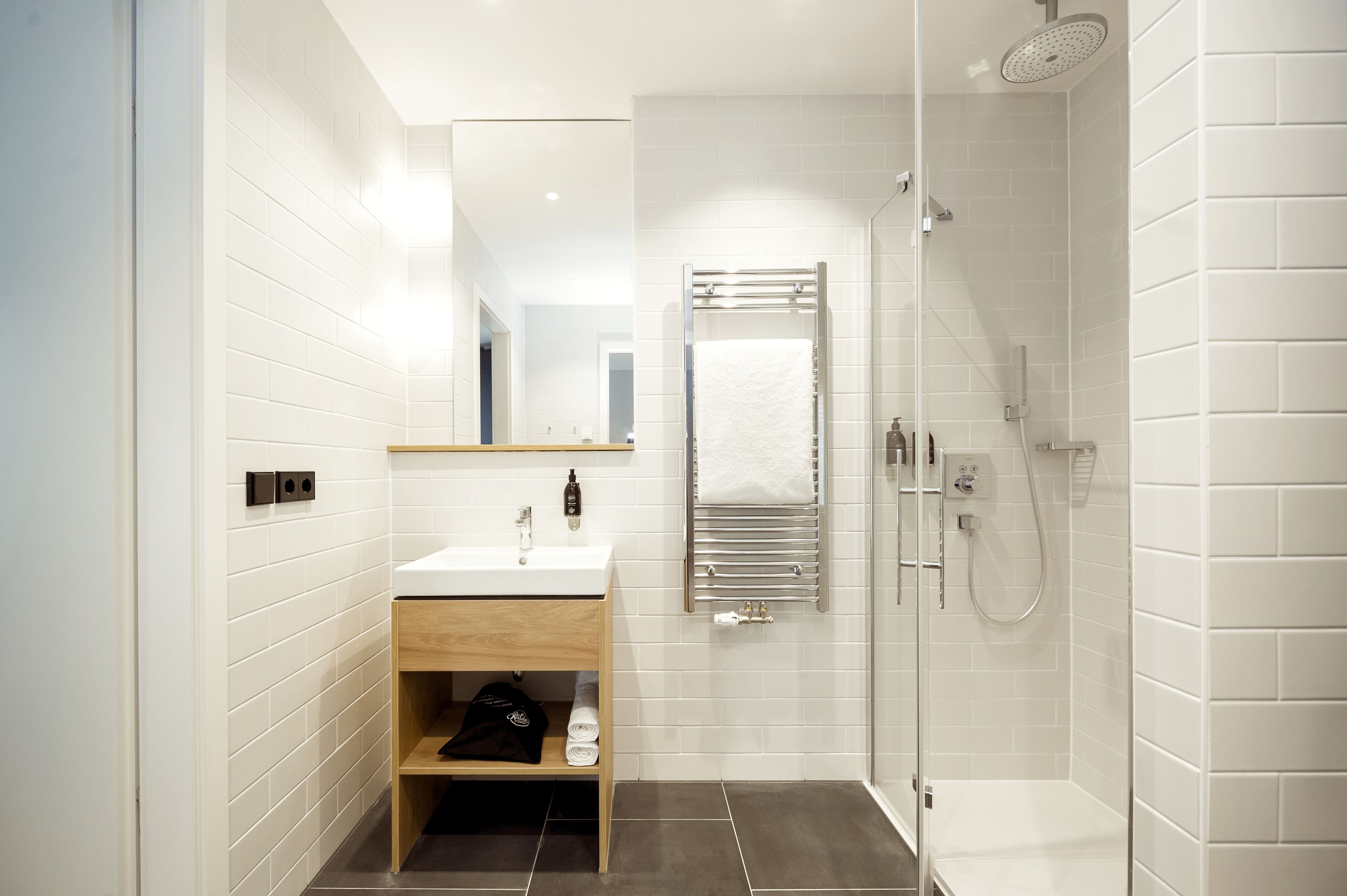 Un baño moderno con pared de azulejos blancos, tocador de madera con lavamanos, cabina de ducha de vidrio y un perchero para toallas.