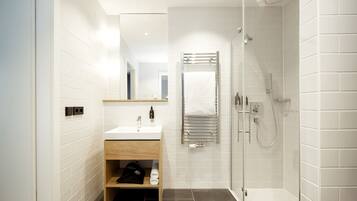 Un baño moderno con pared de azulejos blancos, tocador de madera con lavamanos, cabina de ducha de vidrio y un perchero para toallas.