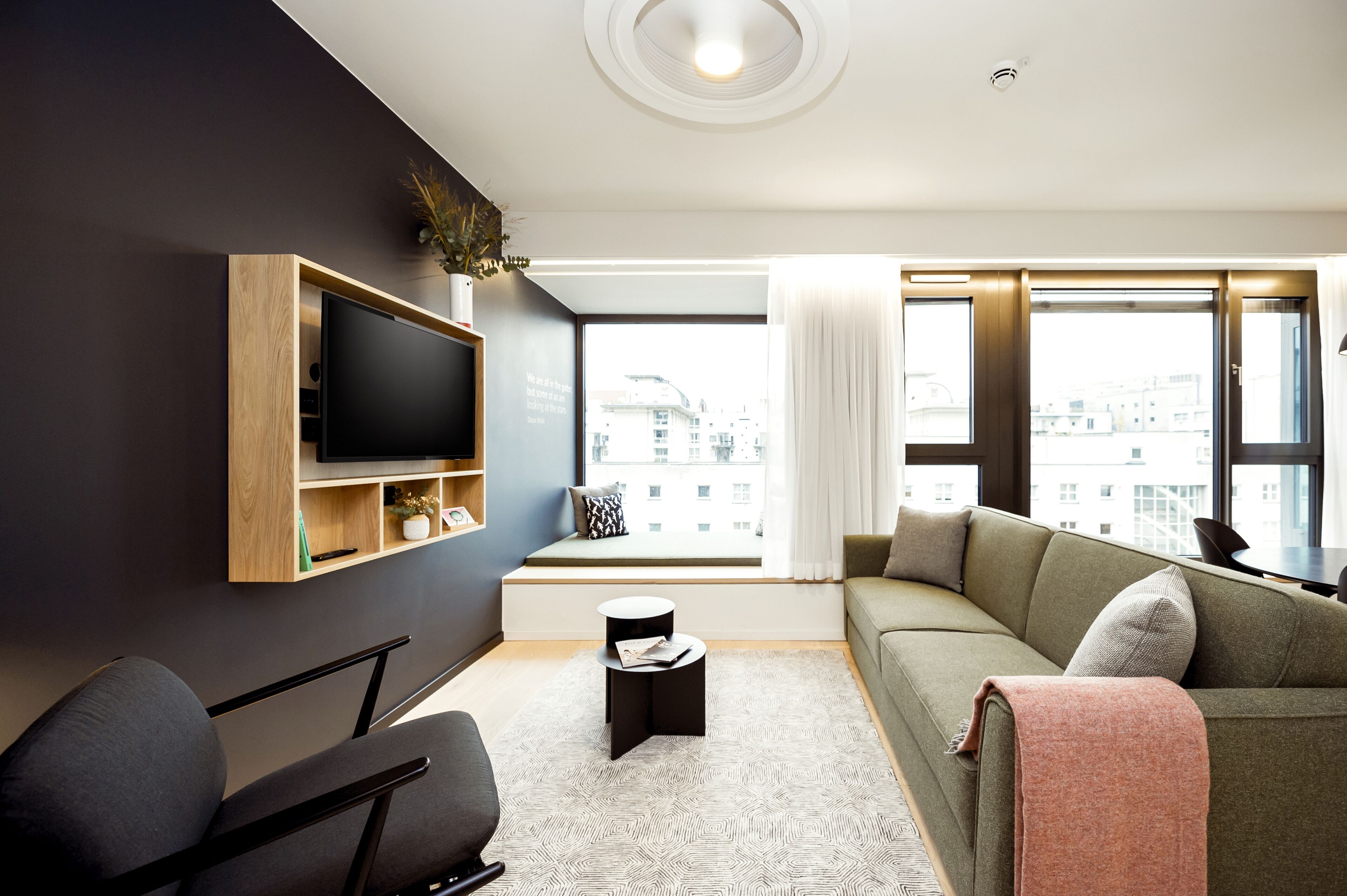 A modern living room with a flat-screen TV mounted on a wooden shelf, a sofa with a throw blanket, a small side table, and a window with a view of buildings.