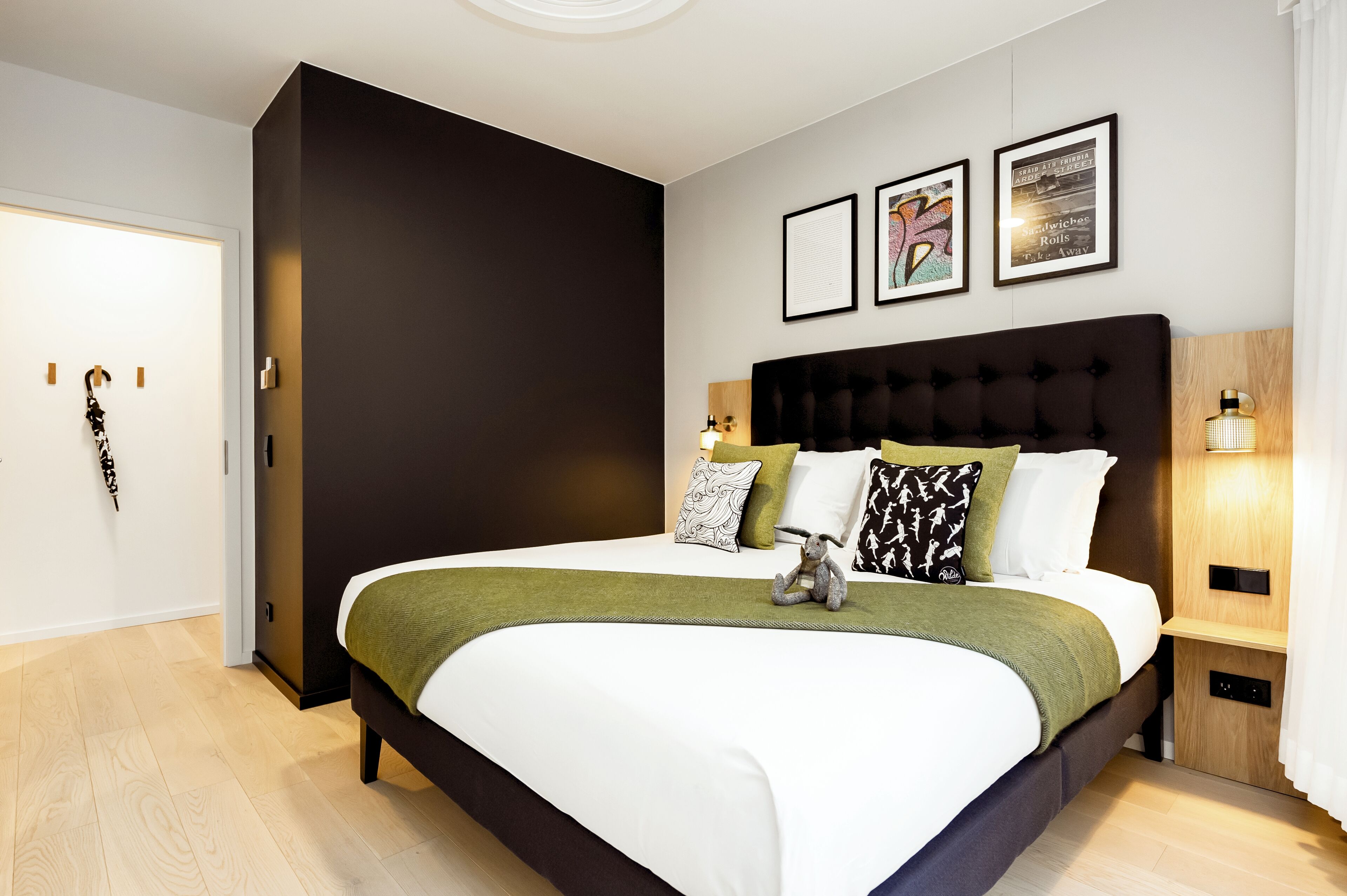 Un dormitorio moderno con una cama grande, cabecera oscura y una manta verde. Hay tres cuadros enmarcados en la pared, una mesita de noche con lámpara y una mesita auxiliar de madera.