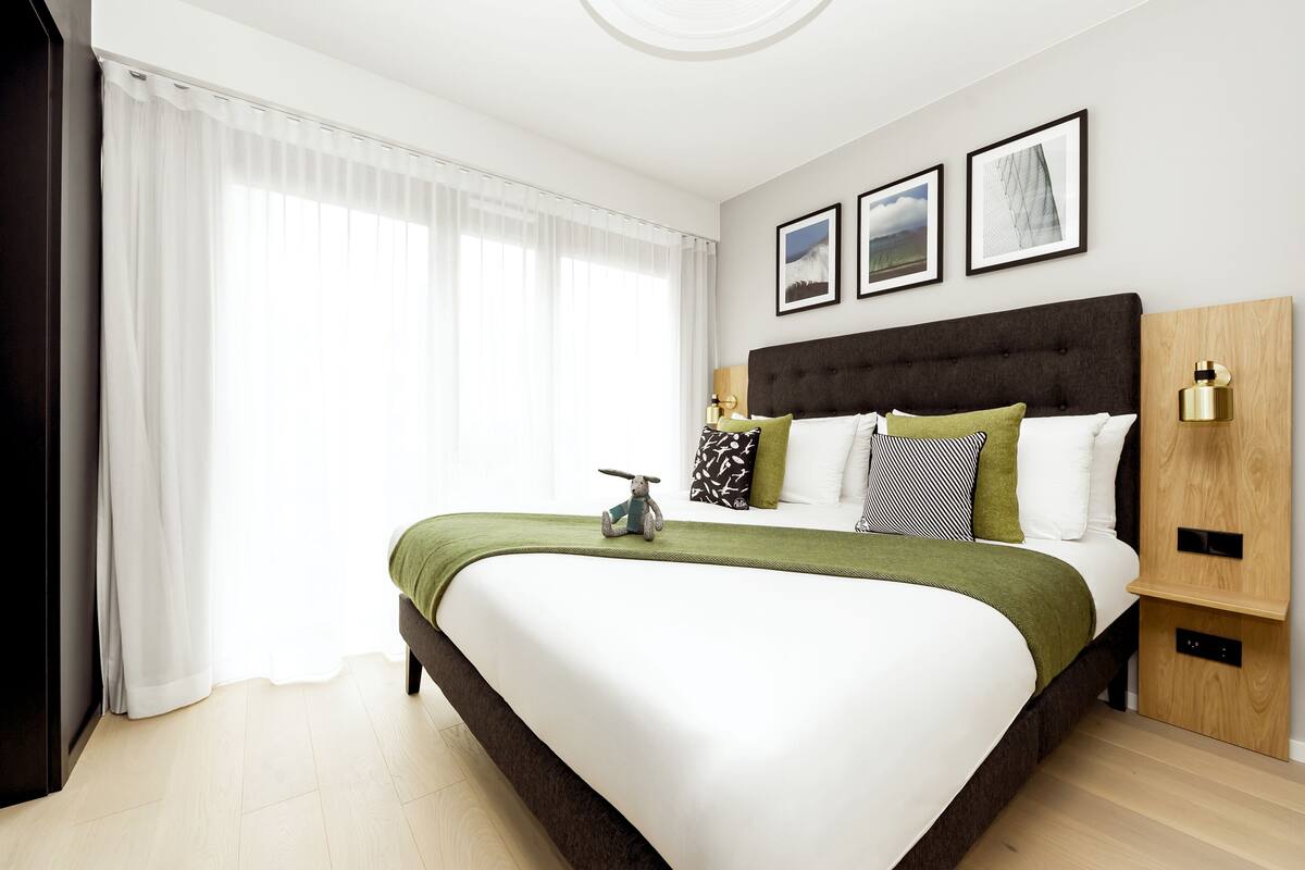 A modern hotel room with a large bed, a wooden headboard, a green throw blanket, and a small toy on the bed.