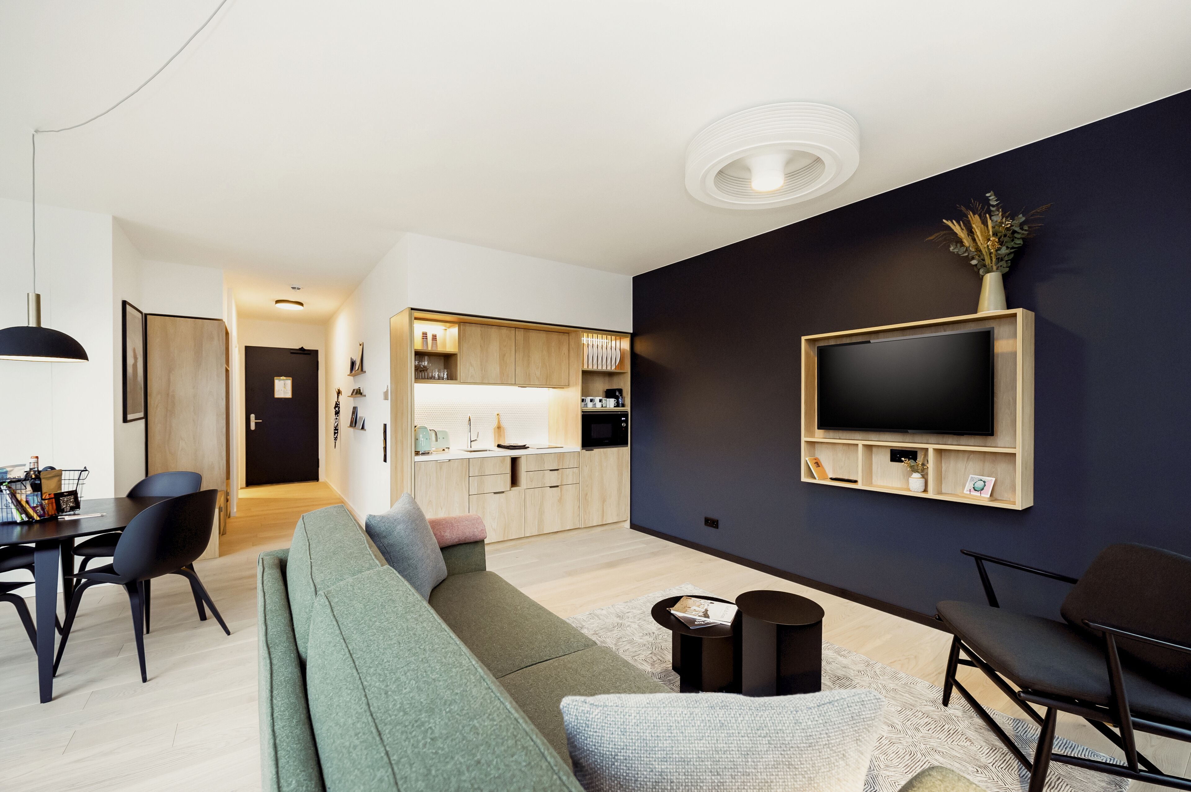 A modern living room with a large flat-screen TV mounted on a dark wall, a green sofa, a dining area with a table and chairs, and a kitchen in the background.