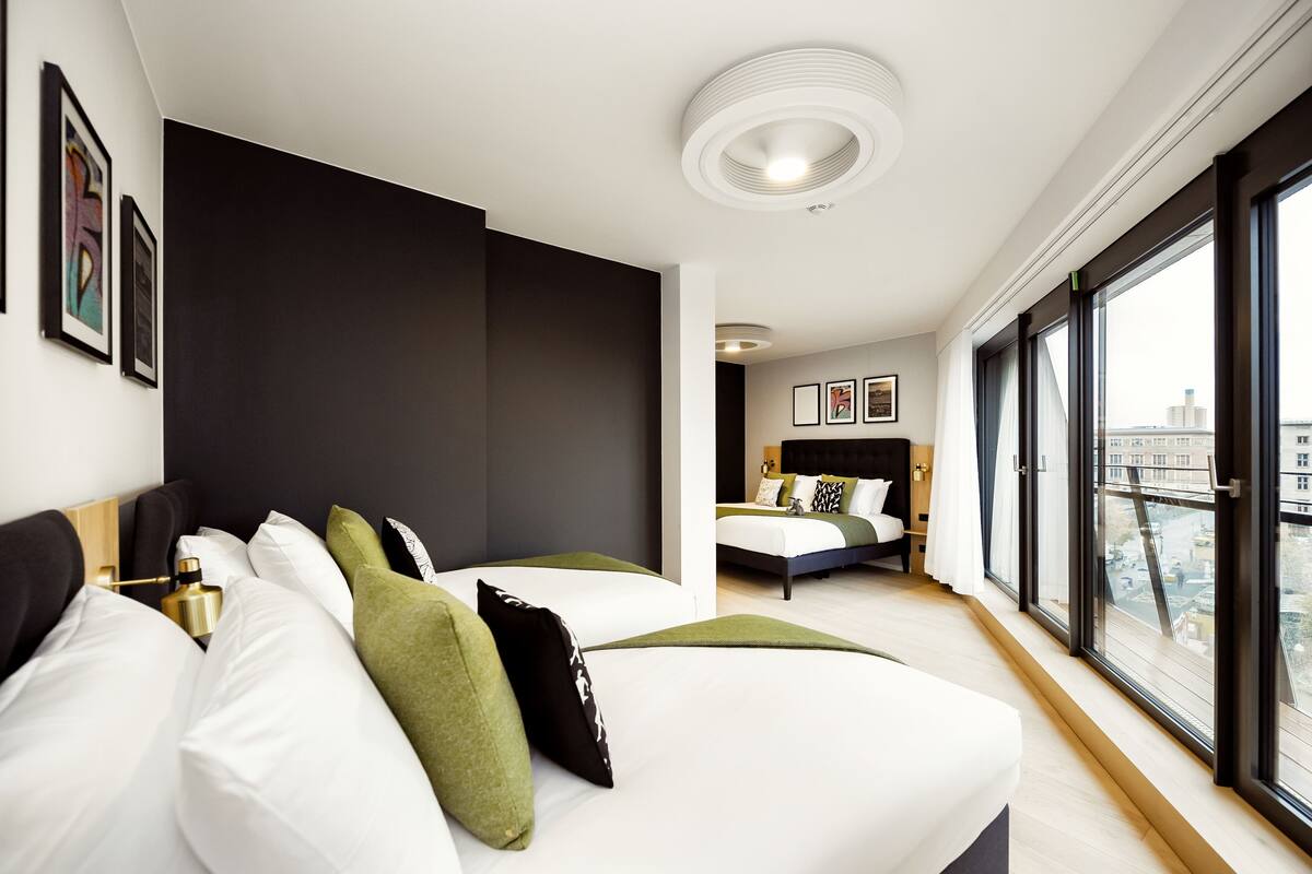 A modern hotel room with a large bed, a smaller bed, and a view of the city through large windows.