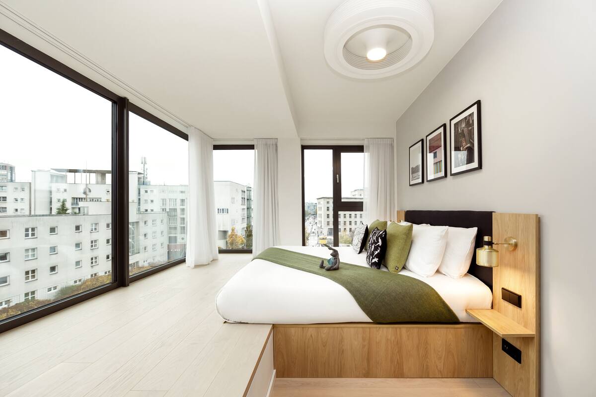 A modern bedroom with a large bed, a wooden bedside table, a nightstand, and a wall-mounted lamp. There are three framed pictures on the wall.