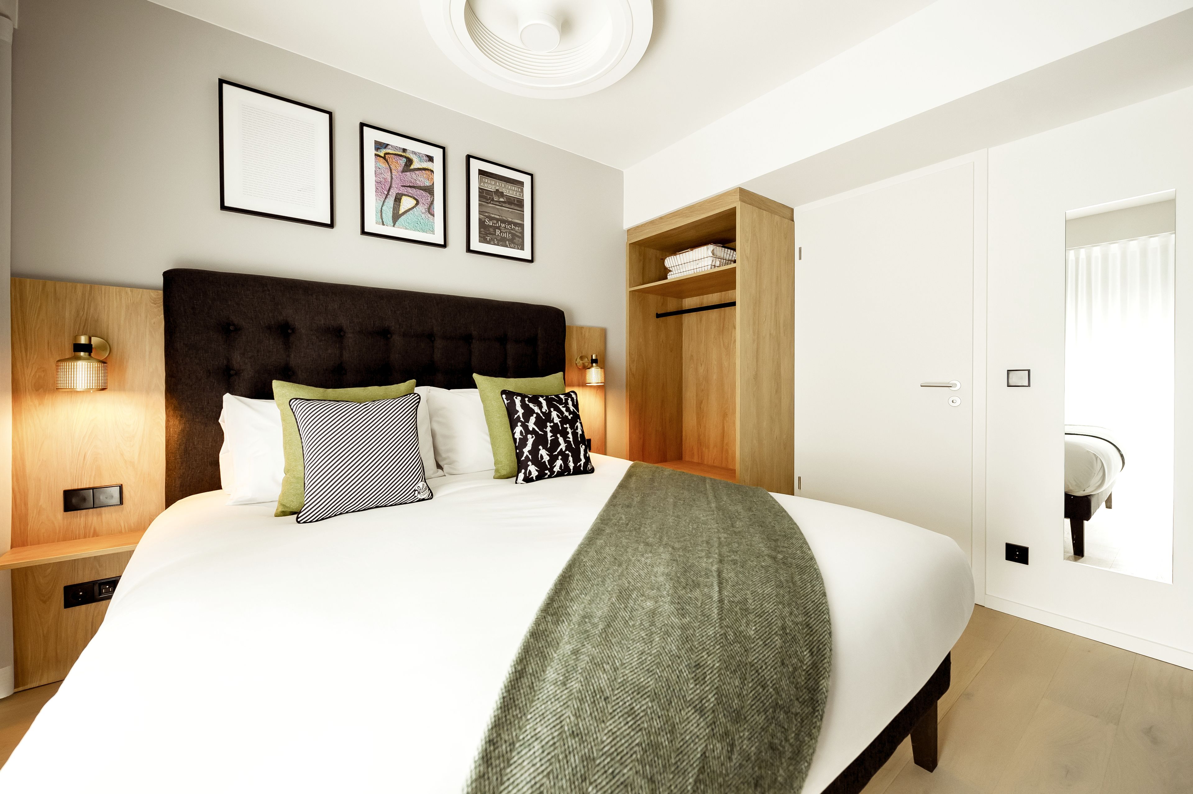 A modern hotel room with a large bed, wooden headboard, and bedside tables with lamps. There are framed pictures on the wall and a wardrobe with a mirror.