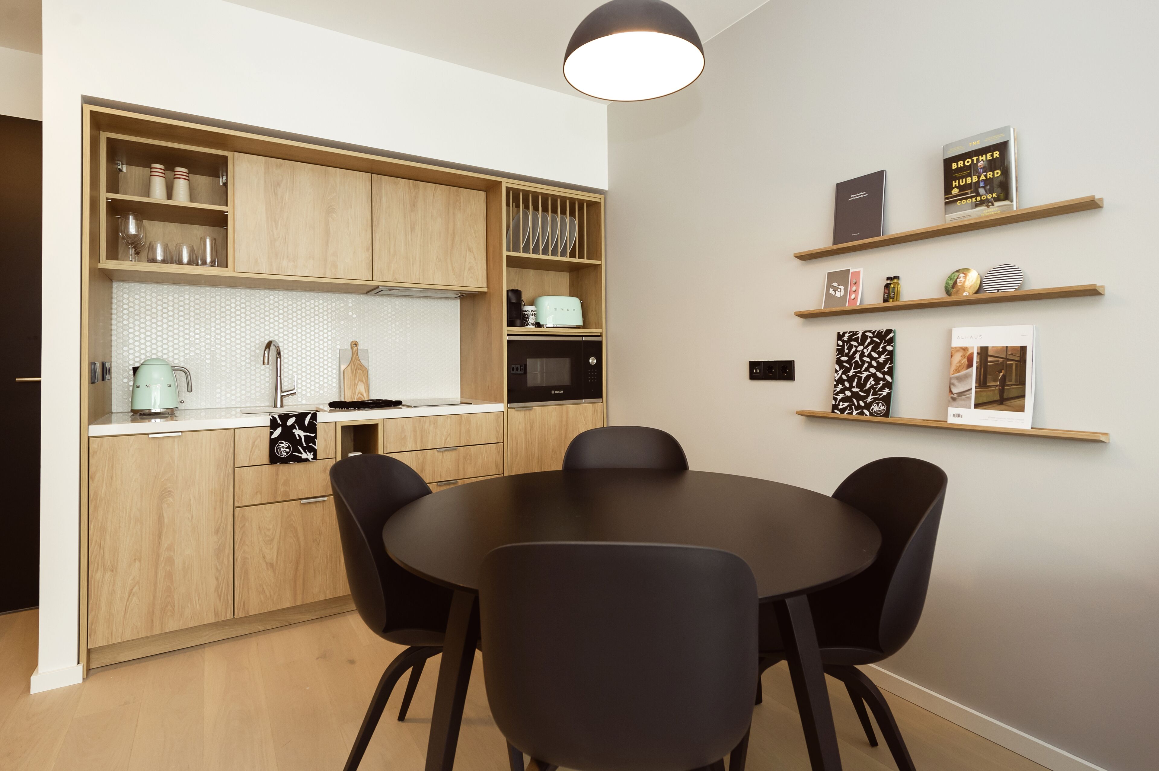 A modern kitchen with wooden cabinets, a round dining table, and black chairs. There are shelves with books and decorative items on the wall.