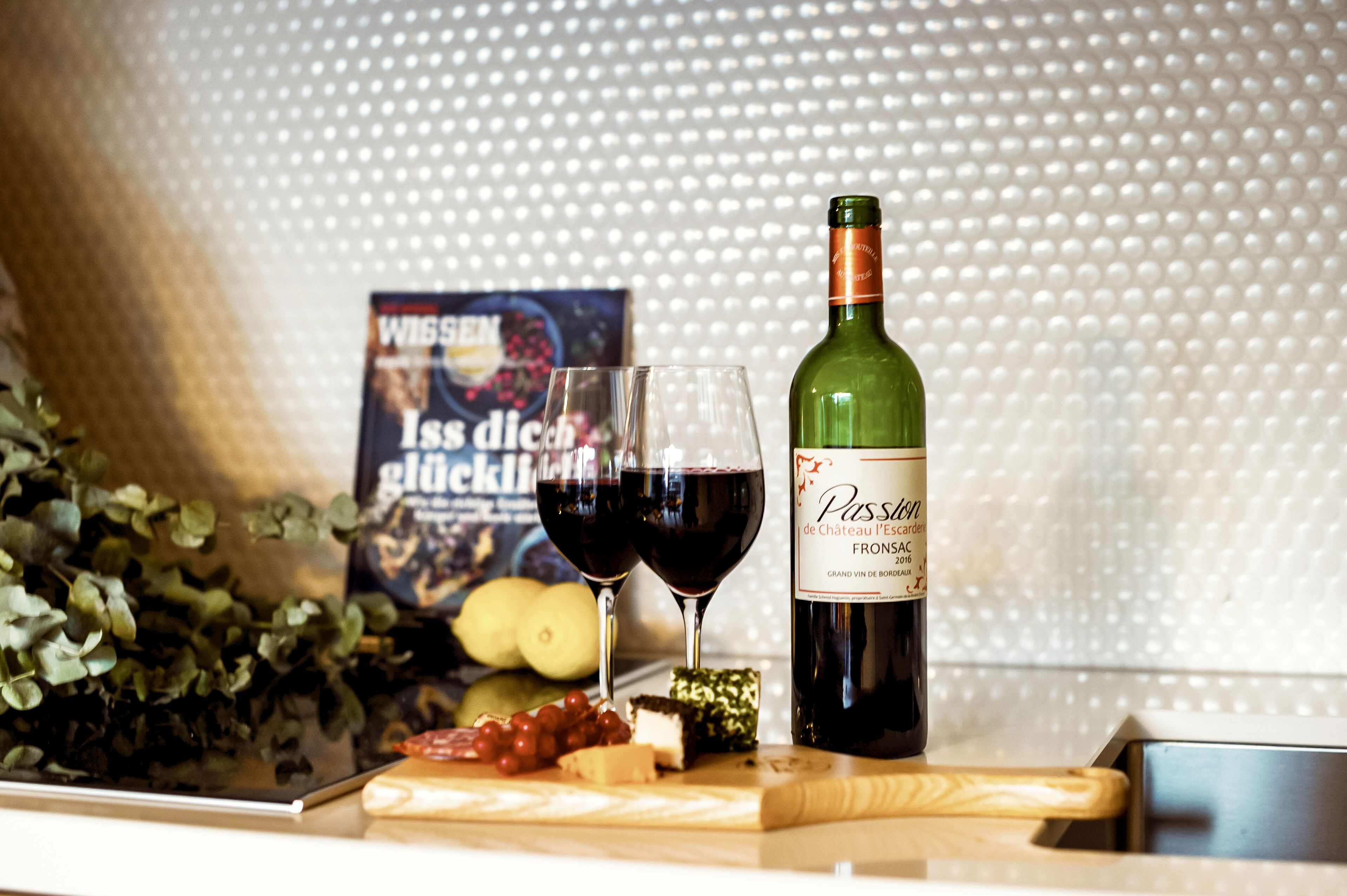 Una botella de vino, dos copas de vino tinto, un libro, queso, uvas y una tabla de cortar sobre una mesa.