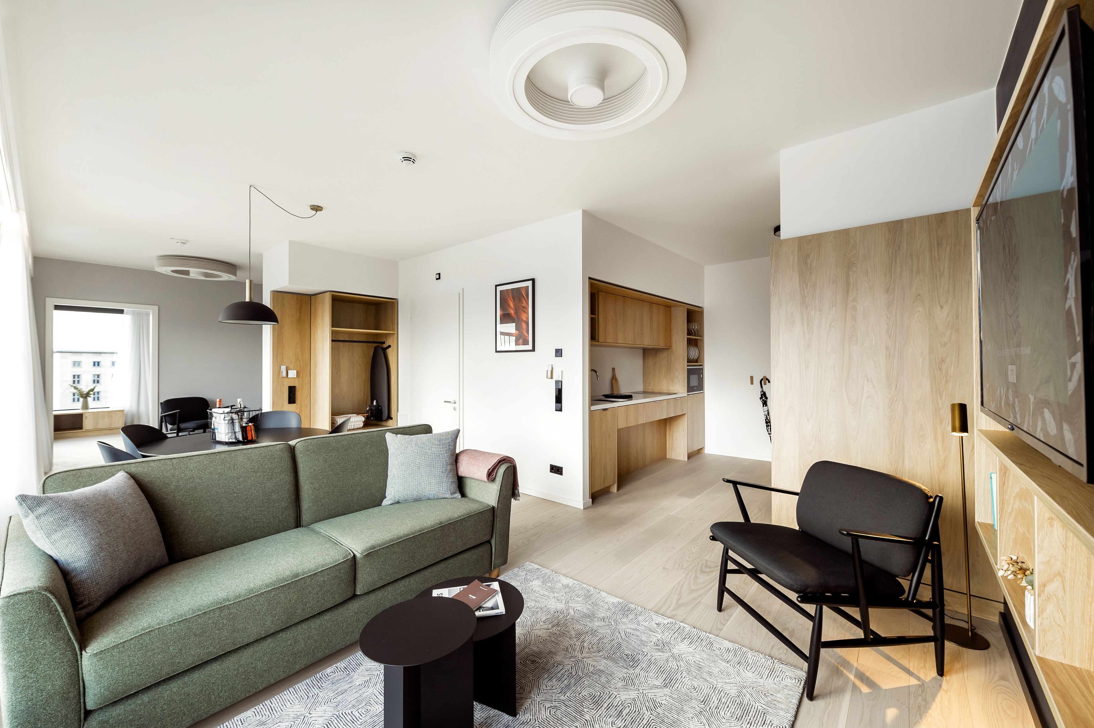 Una sala de estar moderna con un sofá, una mesa de centro y un televisor. Se observa una zona de cocina con armarios de madera y una mesa de comedor.