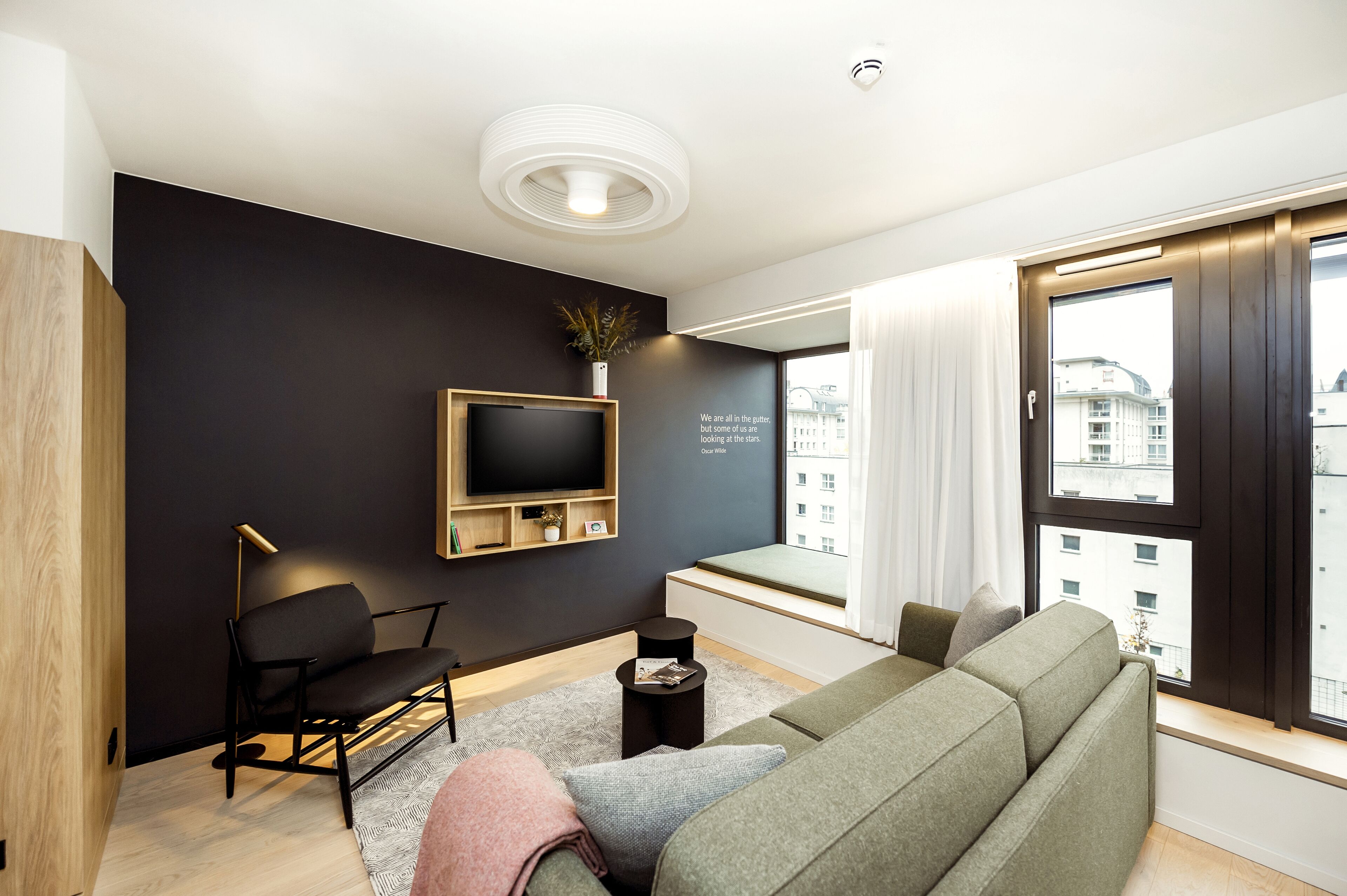 Een moderne woonkamer met een donkere muur, een wandtelevisie, een bank, een stoel en een raam met uitzicht op gebouwen.