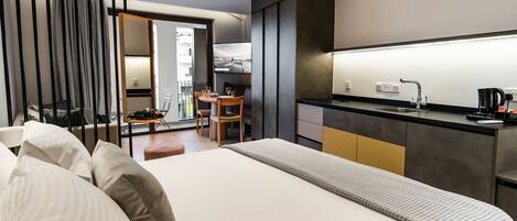 Una habitación de hotel moderna con una cama grande, un área de comedor con mesa y sillas, una cocina con mostrador y electrodomésticos, y un acceso corredizo a un espacio exterior.