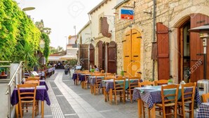 Un café al aire libre con mesas y sillas, edificios de piedra y follaje.