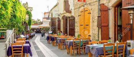 An outdoor café with tables and chairs, stone buildings, and greenery.