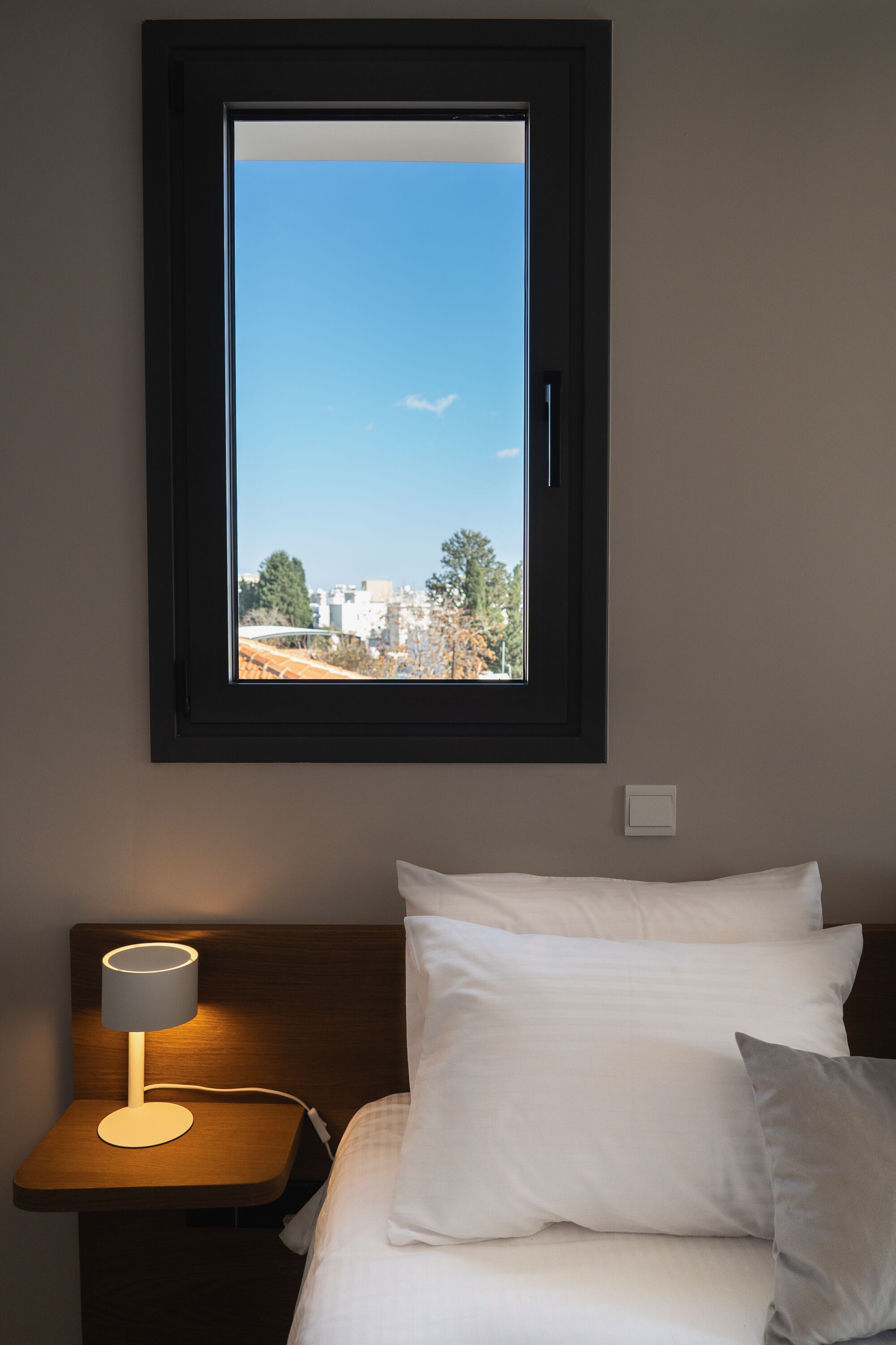 A bedroom with a bed, white pillows, a bedside lamp, and a window with a view of buildings and trees.