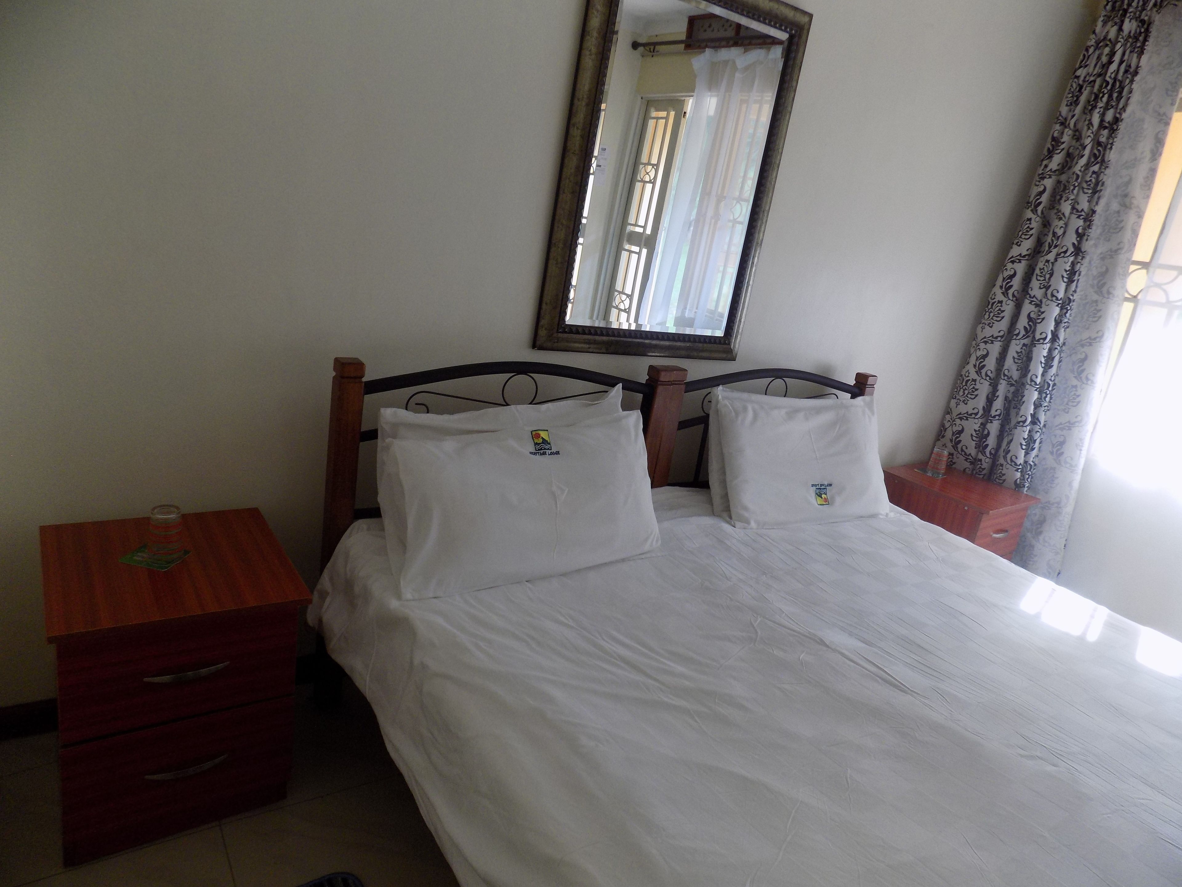 Un dormitorio con cama, dos almohadas, una mesita de noche de madera, un aparador y un espejo en la pared.
