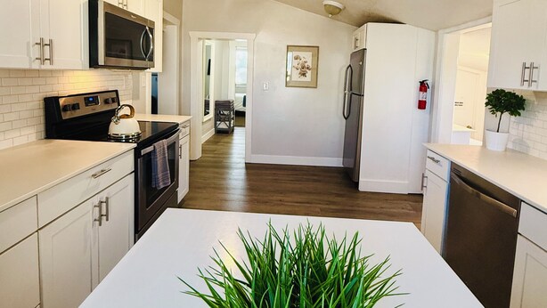 A modern kitchen with white cabinetry, a central island with a green plant, stainless steel appliances, and a wooden floor. - The Cozy Murray Bungalow fast WIFI free parking!  (Murray)