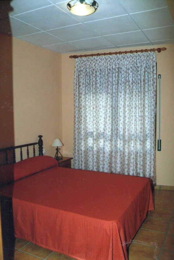 Un dormitorio con cama, mesita de noche, lámpara y cortina.