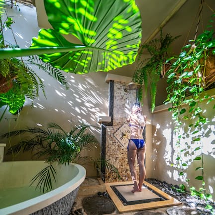 A person in a blue bikini standing in a naturalistic indoor setting with a stone-tiled floor, a large indoor water feature, and lush green foliage.
