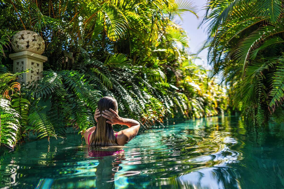A person in a clear water body surrounded by lush greenery and a decorative structure.