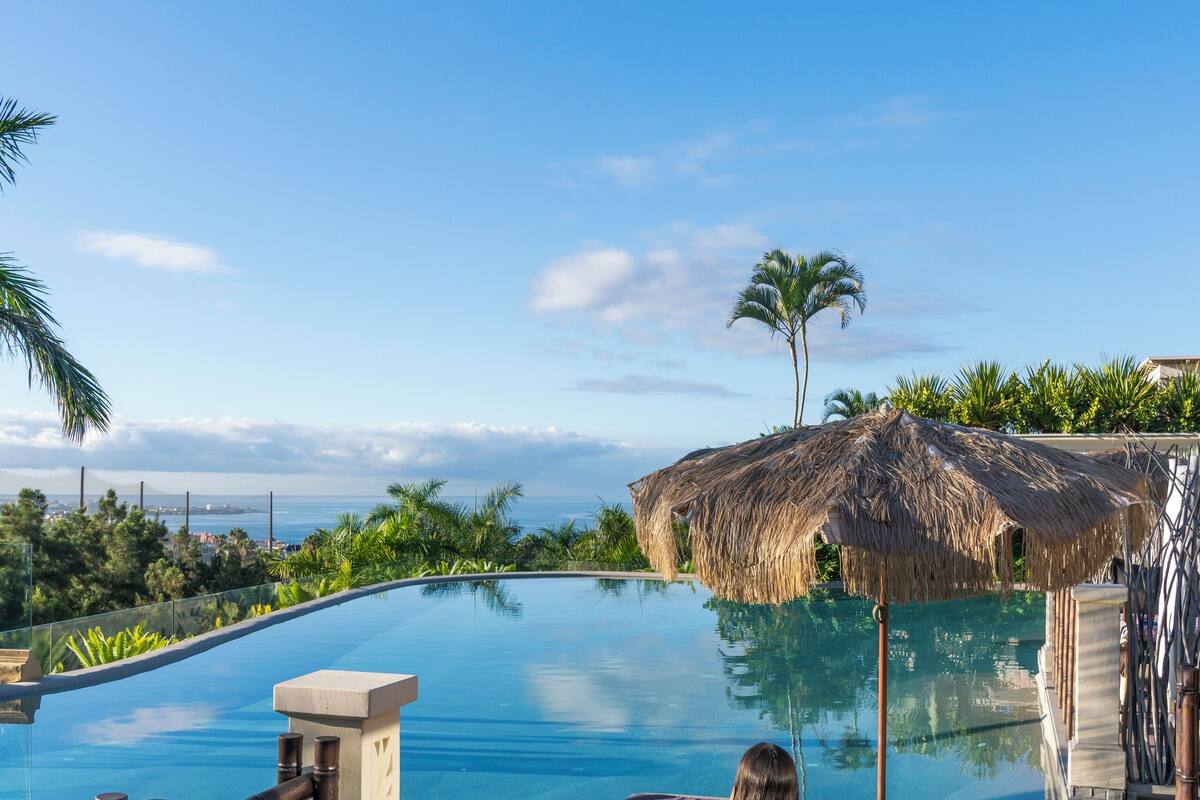 A poolside lounge area with a view of the ocean, a thatched umbrella, and a wooden deck.
