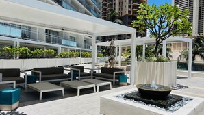 An outdoor seating area with sofas and tables, a water feature, and a covered section with plants. - Ilikai Apt 302 -Newly Renovated October 2025- Studio with  Ocean & Harbor Views Modern Updates (Waikiki)