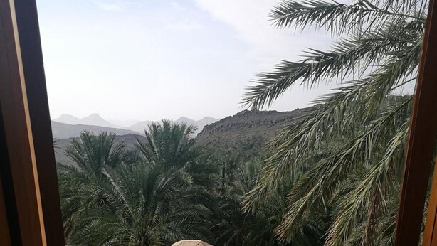 A balcony with a table and chairs, surrounded by palm trees and mountains in the background. - Misfah Old House (Al Hamra)