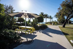 A sunny outdoor area with a picnic table and umbrella, surrounded by greenery and palm trees.