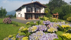 Una casa de dos pisos con balcón, rodeada de exuberante vegetación y flores.