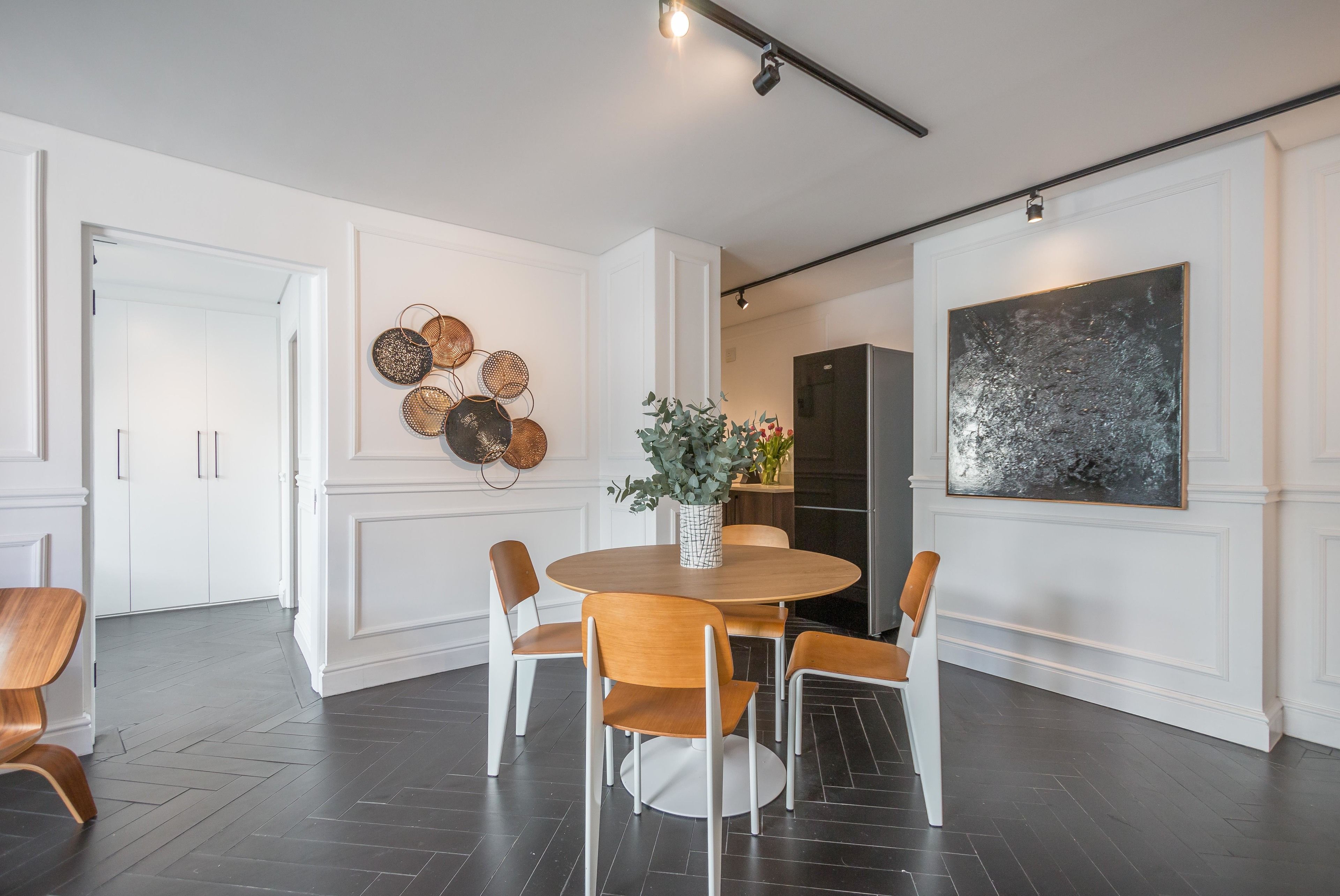 A modern dining area with a round table, four chairs, a vase with flowers, and a large abstract painting on the wall.