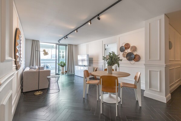 Una sala de estar moderna con sofá, mesa de comedor y sillas. Hay un televisor, una planta y elementos decorativos en la pared.