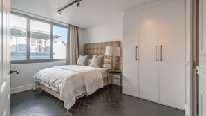 A bedroom with a large bed, a nightstand, a lamp, and a wardrobe. There is a window with curtains and a view of buildings outside. - Greenmarket 701 - Adults Only (Cape Town)