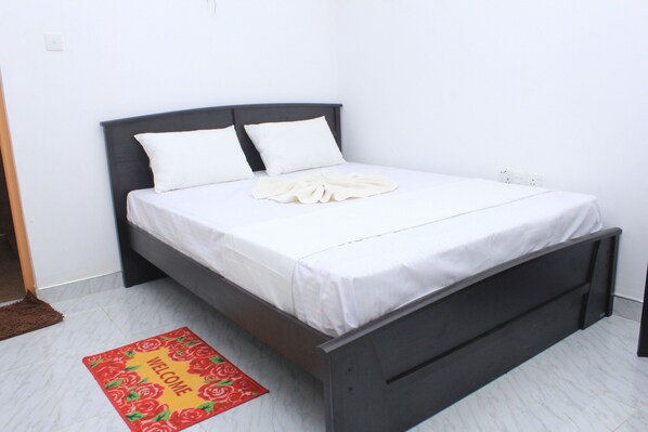 A neatly made bed with white bedding and pillows, a dark headboard, and a welcome mat on the floor. - Nature Night Homestay (Sigiriya)