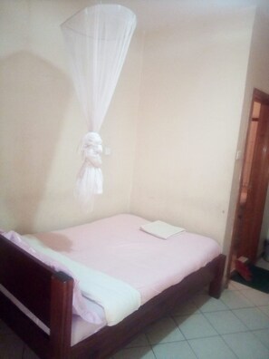 A single bed with a wooden frame and a pink mattress, a white mosquito net, and a white wall.