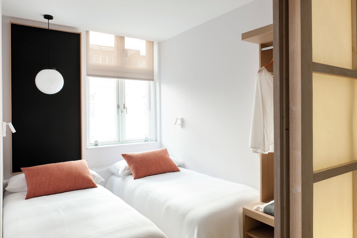 Habitación de hotel con dos camas individuales, una pared de acento negra, una lámpara redonda y una ventana con persianas.