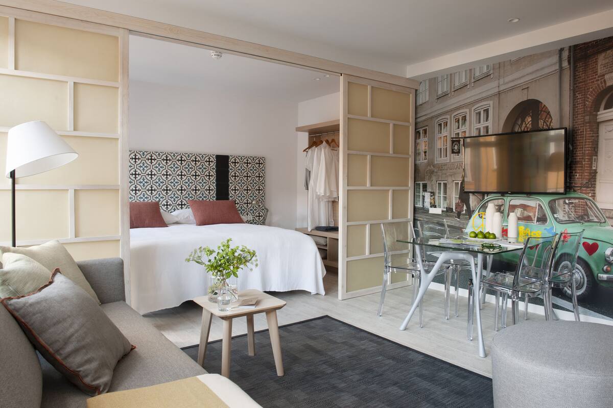 A modern hotel room with a bed, a sofa, a dining table, and a mural of a vintage car on the wall.