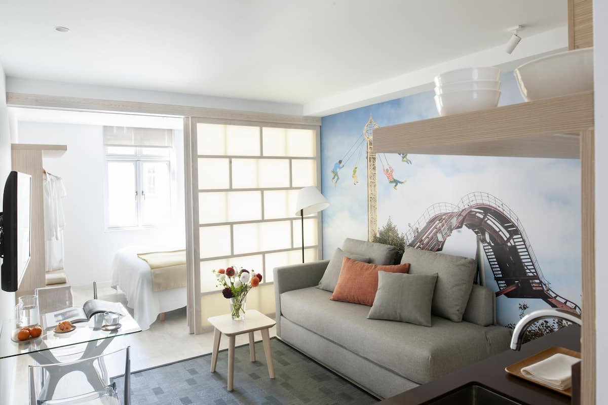 A modern hotel room with a grey sofa, a glass coffee table, a small side table with a vase of flowers, a bed with white linens, and a mural of a roller coaster on the wall.