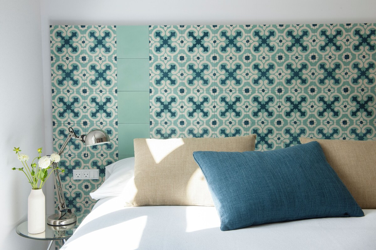Una cama bien hecha con dos almohadas, una lámpara de noche y un jarrón con flores sobre una mesita de noche.