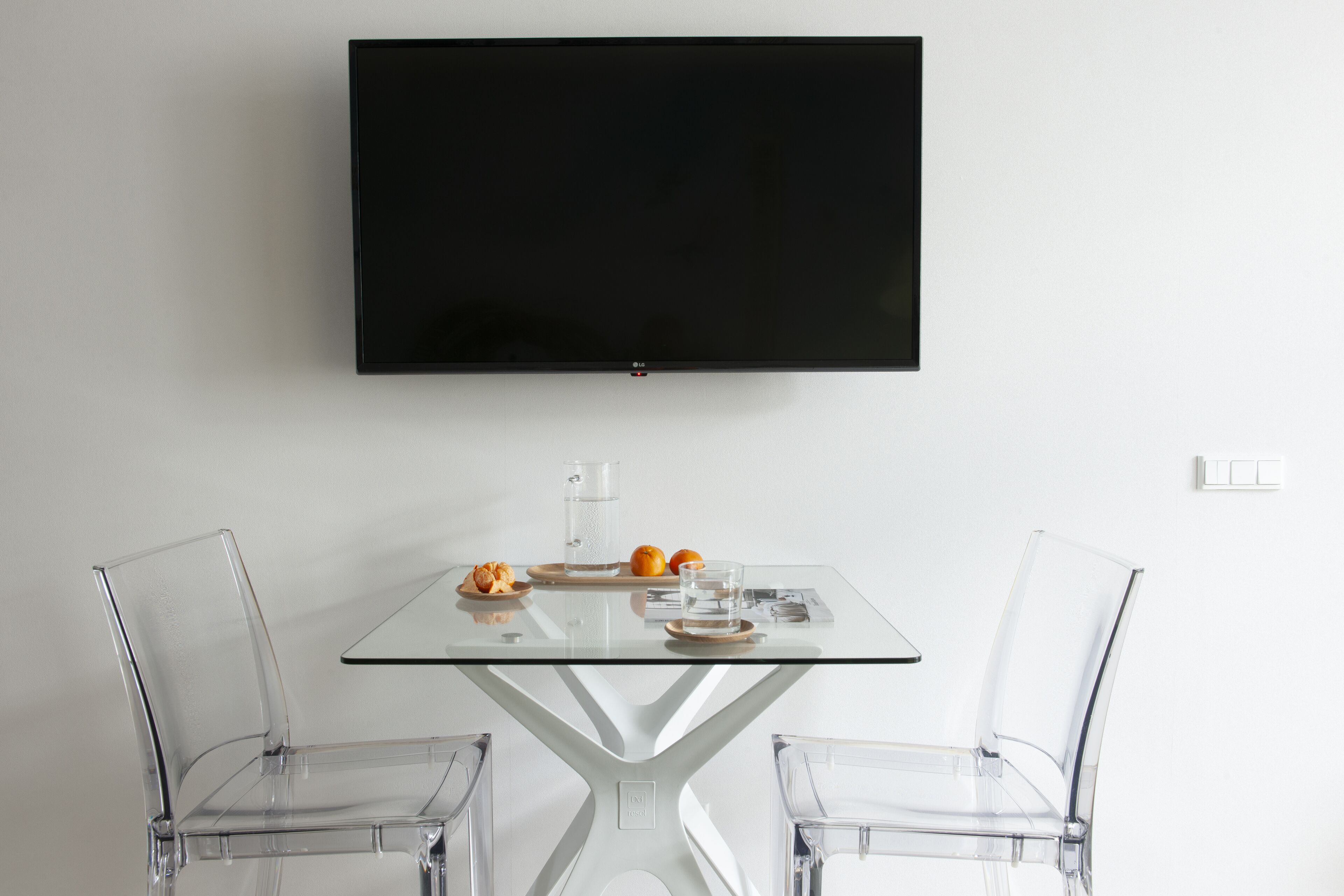 A modern room with a flat-screen TV mounted on the wall, a glass table with two chairs, and a tray with a glass of water, an orange, and some snacks.
