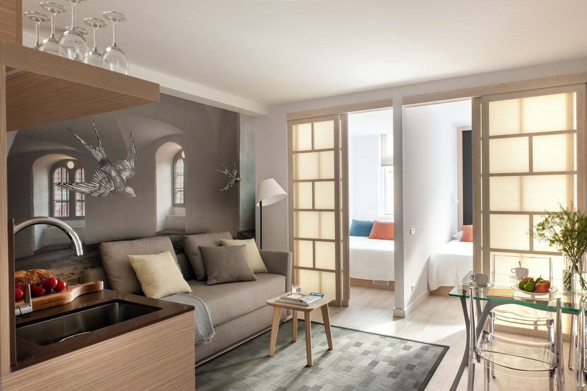 Quarto de hotel moderno com cozinha compacta, sofá, área de refeições e uma grande janela com vista.