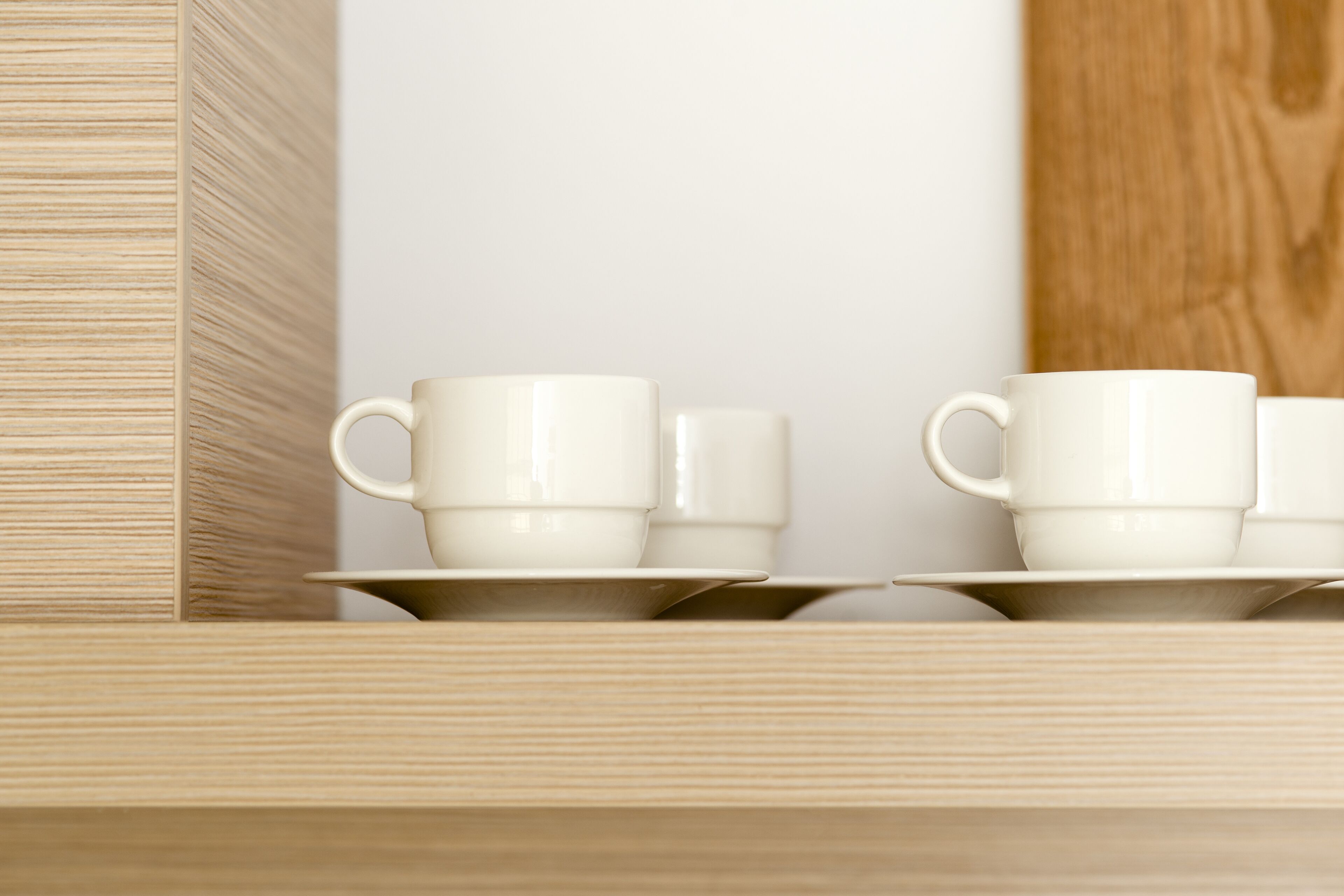 Un ensemble de tasses et de soucoupes en céramique blanche, posées sur une étagère en bois devant un mur blanc.