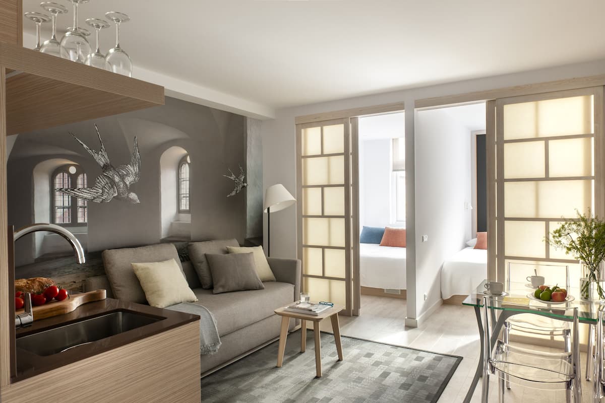 A modern hotel room with a kitchenette, a sofa, a glass dining table, and a bed with a view.
